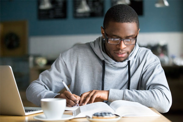 Young african-american male studying with open book, notebook, and laptop on table