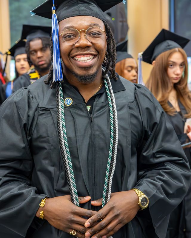 NSCC graduate stands smiling in front of the Lynn Campus building wearing a graduation cap and gown celebrating their achievement after the commencement ceremony